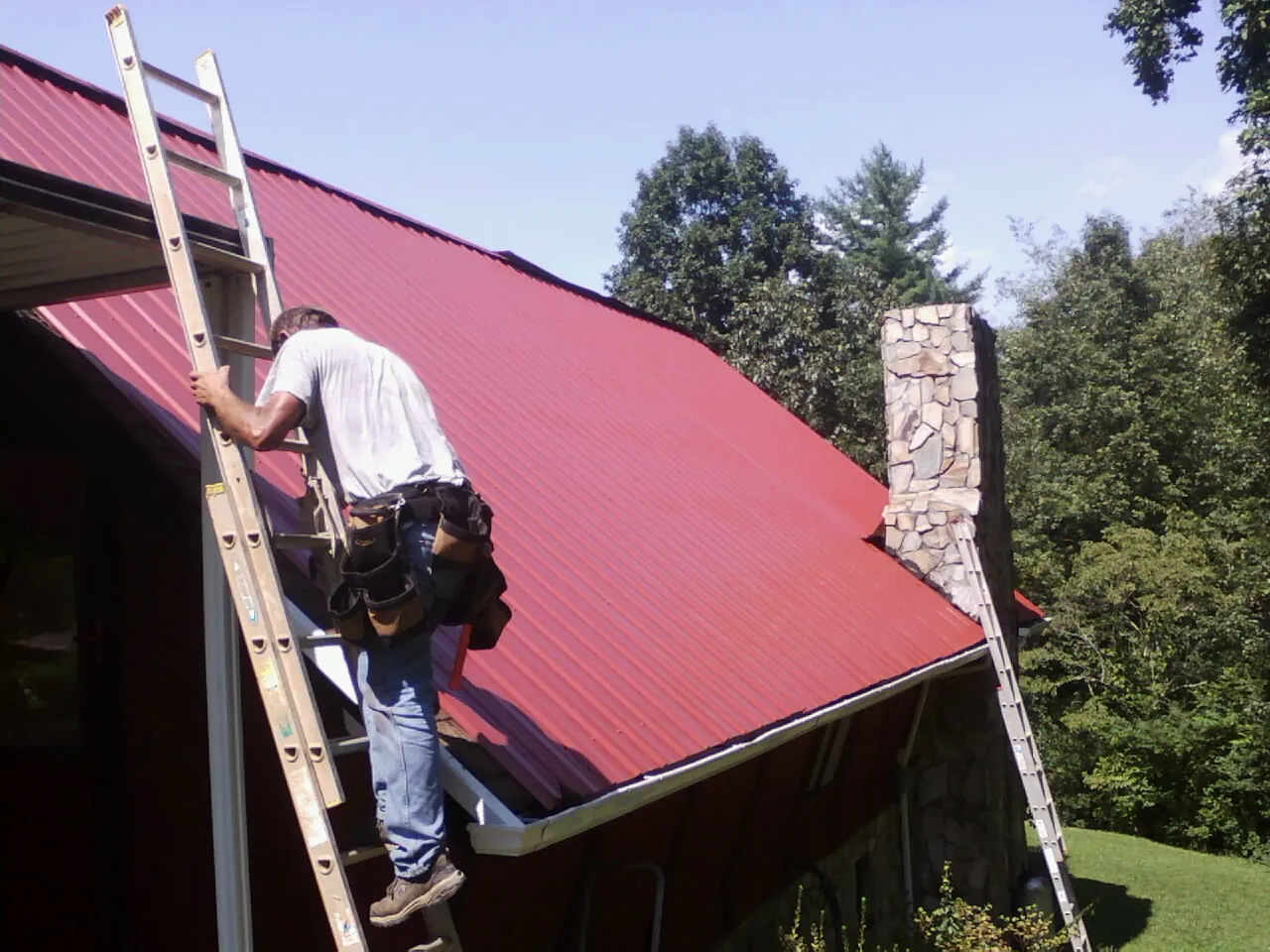 Roofer on a ladder beside a red metal roof and stone chimney — Soffit & Fascia Repair by Roofing Brothers in Reinholds